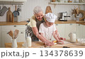Happy family in kitchen. Grandmother and granddaughter child cook in kitchen together. Grandma teaching kid girl roll out dough bake cookies. Household teamwork helping family generations concept 134378639