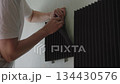 Cropped shot of male hands peeling off dark soundproofing panel from wall, revealing adhesive residue, engaging in room renovation and acoustic treatment removal for interior space. 134430576