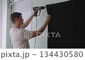 Young man using heat gun to remove old soundproofing panels from wall, undertaking renovation project and home repair, working on sound insulation improvement. Shooting in slow motion. 134430580