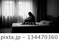 Wide shot of a sad woman silhouette sitting alone on a bed in a dark room backlit by a window 134470360