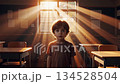 Young caucasian child standing alone in empty classroom with dramatic golden sunlight streaming through window creating magical dust particles and warm cinematic atmosphere 134528504