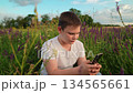Child sits on grass in park uses smartphone. Boy spends digital leisure time in meadow. Happy boy uses gadget outdoors. Technology in boy hands, meadow flowers. Teenage boy plays on smartphone nature 134565661