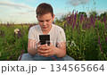 Teenager boy playing on smartphone nature. Child sitting on grass watching smartphone. Happy boy child using gadget outdoors. Technology in boy hands lawn with flowers. Boy digital leisure on meadow 134565664