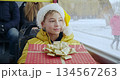 Front view of small boy sitting on bus, smiling. Kid in Christmas hat holding gift box, looking forward, glad, happy. Concept of New Year, Christmas and winter vacations. 134567263