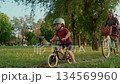 Little boy wearing a helmet rides a bicycle in a green park, with his mother  134569960