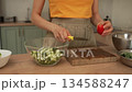 Close-up of female hands cutting fresh vegetables on a wooden board in a modern kitchen. The woman is chopping a ripe red tomato for a healthy, nutritious salad with sliced cucumbers 134588247