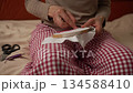 Woman in plaid pajamas is seated on a bed, engaged in embroidery at home. The camera zooms in on her expert hands as she meticulously stitches a fabric held in a wooden embroidery hoop 134588410