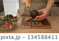 Woman's hands cutting a ripe avocado on a wooden board to prepare a healthy homemade vegetable salad in a glass bowl, showcasing a nutritious meal preparation and dieting concept 134588411