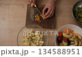 Top-down view of female hands slicing mandarin orange wedges with a knife on a wooden cutting board, preparing a healthy and fresh fruit salad in the kitchen for a nutritious meal 134588951