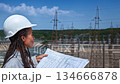 female engineer on the observation deck of a huge hydroelectric power station holding a drawing of the construction project of an engineering structure 134666878