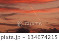 An airplane flies through a colorful sunset sky. Contrails are visible against the vibrant clouds, creating a striking contrast with the warm hues. 134674215