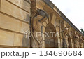 Series of sandstone sculptures supporting arched wall with repeating figures beside stone stairs and green trees. 134690684