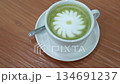 A warm cup of matcha latte rests on a white saucer atop a wooden table, with white foam forming a simple floral design in the center. 134691237