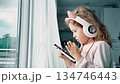 Young child using smartphone while wearing headphones at home near window 134746443