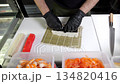 A chef wearing gloves prepares sushi rolls with layered fillings of salmon, avocado, tuna and eel on a bamboo mat in a professional kitchen. Asian cuisine, cooking process. Top view.  134820416