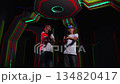 A group of cheerful teenagers with laser guns are having fun in a dark laser tag arena, a boy and a girl are standing with guns in their hands against a wall with neon stripes. 134820417