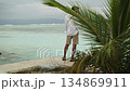 A young man in light clothes with phones in his hands stands on a sandy beach under palm trees. Against the background of the waves of the sea and a cloudy sky. Maldives. 134869911