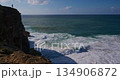 Person Stands Atop a Rugged Cliff, Gazing at the Vast Blue Ocean and Crashing Waves 134906872