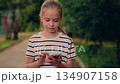 young girl making phone call in nature, girl answering call in peaceful park, small child speaking on cell in nature, child chatting on mobile on sunny path, cheerful child holding phone in park 134907158