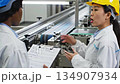 Two professionals in safety gear discuss production process at conveyor belt, showcasing teamwork and communication, camera pans and zooms in 134907934