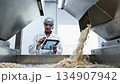 Female food technician monitors production process, observing ingredient flow while using tablet, showcasing quality control in manufacturing environment 134907942
