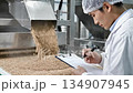 Technician observes grain pouring from a machine while taking notes, showcasing quality control in a production environment with copy space 134907945