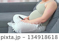 Pregnant woman sitting on a couch using her smartphone in a relaxed home setting. A calm everyday moment highlighting modern maternity lifestyle, comfort, and digital communication during pregnancy 134918618