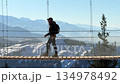 A side view of a backpacker crossing a long suspension bridge on a sunny winter day against a stunning alpine backdrop 134978492