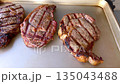 A person skillfully uses tongs to flip sizzling steaks on a charcoal grill, with smoky flavors enhancing the juicy meat, accompanied by halved onions grilling alongside. 135043488