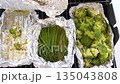 A closeup shows sliced carrots cooking inside a foil packet on an outdoor grill. The bright orange color contrasts against the silver foil and dark grill surface. 135043808
