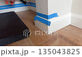 Caulking gun used to seal the gap between white baseboard and wall with blue tape applied. Shows DIY home improvement technique. 135043825