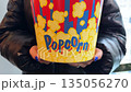 Round cardboard popcorn bucket with bright design. Person holds popcorn before going to cinema. Movie night with favorite snack. Person holds bucket of popcorn in front 135056270