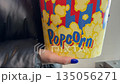 Large bucket of popcorn in hands of girl with blue manicure. Bright nails on hands of girl holding popcorn snack for cinema. Concept of going to movies with popcorn 135056271
