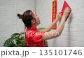 Happy young Asian woman wearing traditional red cheongsam dress decorating home with lucky couplets banner for Chinese New Year festival celebration Happy young Asian woman wearing traditional red cheongsam dress decorating home with lucky couplets banner for Chinese New Year festival celebration 135101746