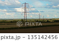 Multiple pylons stretching across farmland transmitting electricity through countryside. Power lines carrying energy transmission. Series of lattice towers extending over fields transferring electric 135142456