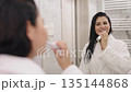 Happy smiling, Woman Mirror, Bathroom Routine. In bathroom, charming lady brushes teeth post-shower, looking at mirror while attired in white silky bathrobe 135144868