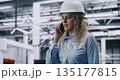 Female Engineer Wearing Hard Hat Walks Across Production Workshop and Speaks on Mobile Phone 135177815