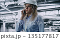 Professional Woman With Curly Blonde Hair in Protective Helmet Moves Through Manufacturing Plant 135177817