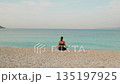 Woman meditating on pebble beach facing calm turquoise sea during peaceful morning 135197925