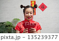 Happy young Asian woman in traditional red Cheongsam dress holding Ang Pao red envelopes for Chinese New Year celebration against white brick background Happy young Asian woman in traditional red Cheongsam dress holding Ang Pao red envelopes for Chinese New Year celebration against white brick background 135239377