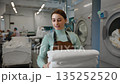 Walking woman worker in an apron holds fresh towels in a laundromat 135252520