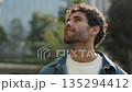 Portrait of happy Hispanic man in casual attire looks upward in city park. Smiling Latin American guy with beard gazes at skyscrapers in summer urban district. Travel destination 135294412