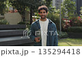 Portrait of young Hispanic man holds digital tablet under arm and smiles in city park. Happy Latin male student in denim jacket with mobile device stands in spring downtown garden 135294418