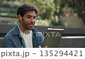 Portrait of bearded Hispanic man talks on video call via digital tablet in park. Happy Latin male discusses plans with girlfriend on online chat via mobile device in urban garden 135294421
