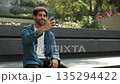 Portrait of happy Hispanic man shows V-gesture on video call by smartphone in city park. Joyful Latin male in casual attire greets friend on online chat via mobile phone on street 135294422