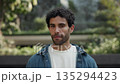 Portrait of curly haired Hispanic man in denim clothes smiles as stands in green park. Happy Latin male with three-day-stubble sends positive vibes resting in spring urban garden 135294423
