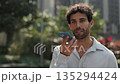 Portrait of Hispanic man in shirt records voice message on smartphone as stands in summer urban park. Bearded Latin male talks on mobile phone with hands free mode slow motion 135294424
