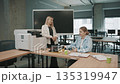 Colleagues discuss office projects while using printer and reviewing documents in a modern workspace 135319947
