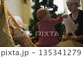 mother persuades baby girl put bright shiny white New Year's hat like hers child quickly takes head asks put, then points finge camera. background Christmas tree waiting decoration with toys and 135341279