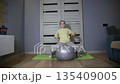 Young Caucasian girl performs gymnastics exercise on fitness ball indoors. Girl practices physical activity at home for health development. 135409005